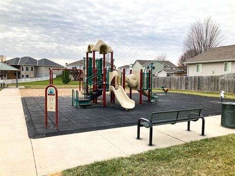 This playground area has a multi level climbing structure with slides and spring riders on a rubber tiled area, next to it, on sand, is a swing set with both belt and bucket swings. There is a paved path around the perimeter. 