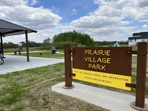 A park sign for Prairie Village Park with a shelter and playfield in the background