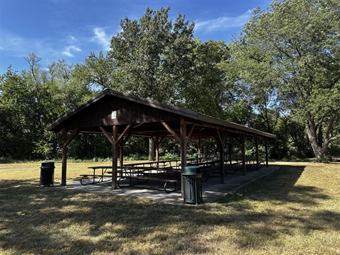 A covered shelter on grass with trees in the background and picnic tables in under the wooden roof.